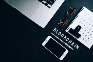 Smartphone, pen, glasses, calendar and laptop on a table, alongside letters spelling 'Blockchain' 